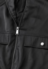 Veste zippée noire en tissu lisse, avec une fermeture éclair argentée, deux poches plaquées et un détail de bouton-pression métallique.