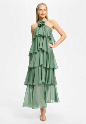 Femme blonde portant une robe en chiffon verte sans manches, à plusieurs couches, avec un volant à l'avant et un décolleté floral, posant avec une main sur la hanche.