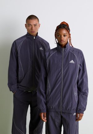 Two models wearing matching dark purple Adidas track jackets and pants, standing against a plain white background.