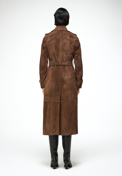 Persoon van achteren gezien die een lange bruine suède trenchcoat met ceintuur draagt en zwarte, kniehoge laarzen met hakken, tegen een witte achtergrond.