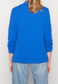 Kungablå, skräddarsydd kavaj med rynkade trekvartslånga ärmar, mjukt tyg, spetskrage och rak nederkant. Bärs tillsammans med ljusblå byxor.