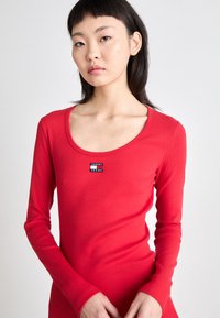 Raudona ilgomis rankovėmis palaidinė, pagaminta iš suspausto audinio, su apvaliu kaklu ir stačiakampiu Tommy Jeans logotipo pleistru ant krūtinės.