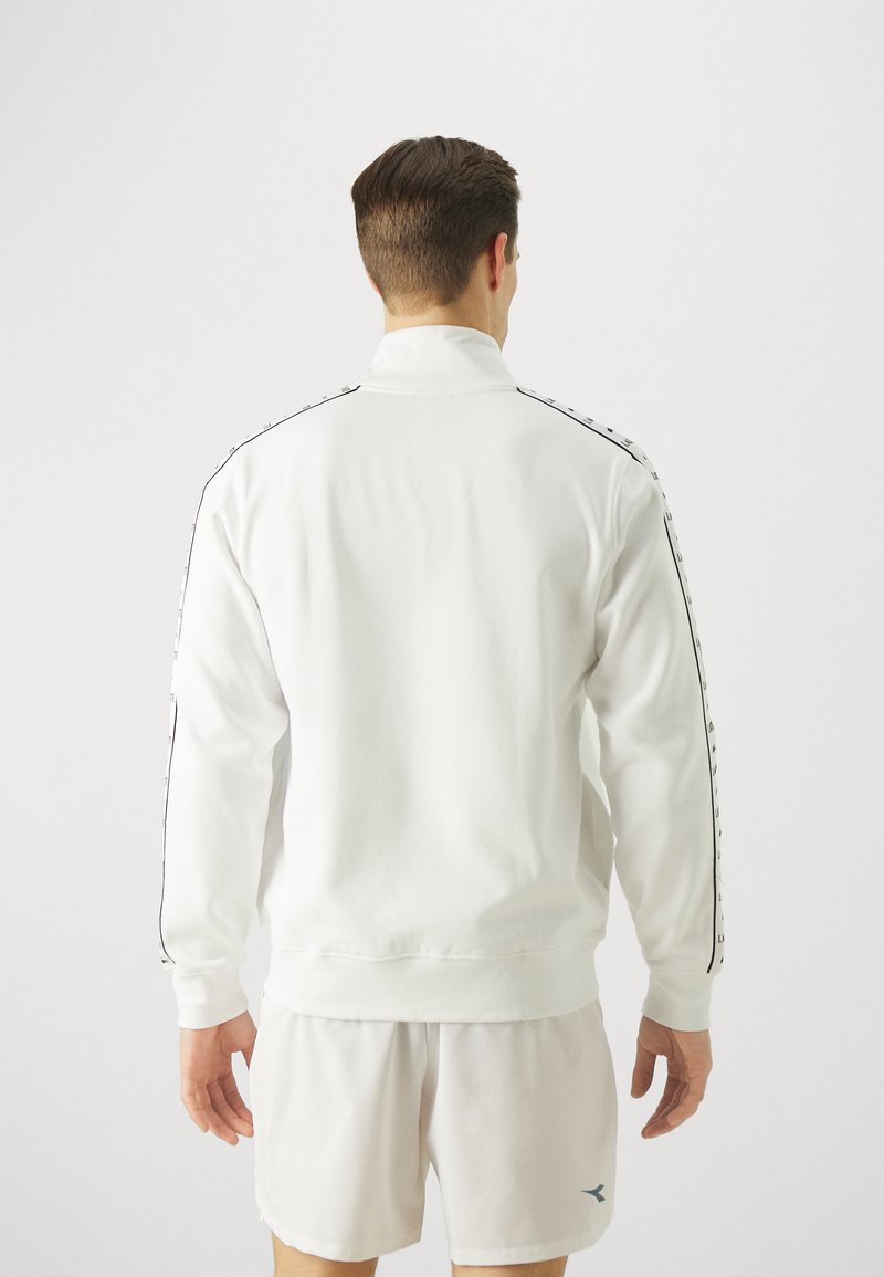 Witte sweatshirt met een hoge kraag, gladde textuur en zwarte zijstrepen met kleine logo's. Gecombineerd met lichte shorts.