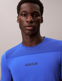 Camisa atlética azul com uma base texturizada, apresenta o logótipo "CALVIN KLEIN" em preto. Material suave e um design ajustado.