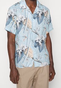 Camicia a maniche corte con bottoni, caratterizzata da uno sfondo azzurro e una stampa botanica in bianco, verde e tonalità pesca. Vestibilità rilassata con colletto a camicia.