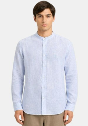 Man die een lichtblauw overhemd met mandarijnkraag en knopen draagt met lange mouwen en beige broek, staand tegen een witte achtergrond.