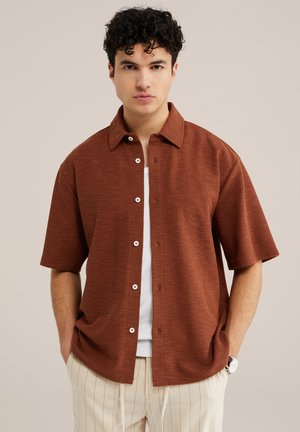 Mladý muž s kučeravými vlasmi, oblečený v neupravených krátkych košeliach hrdzavohnedej farby cez biely top a krémové nohavice s jemnými prúžkami, ruky v vreckách.