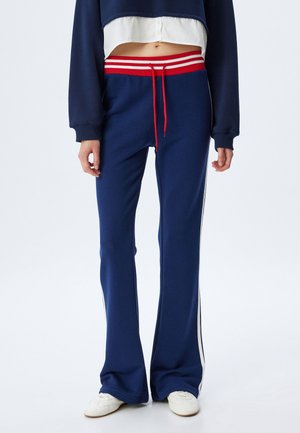 Mørkeblå udvidede sweatpants med en rød- og hvidstripet talje, der har hvide striber på siden og en snor. Glat stoftekstur.