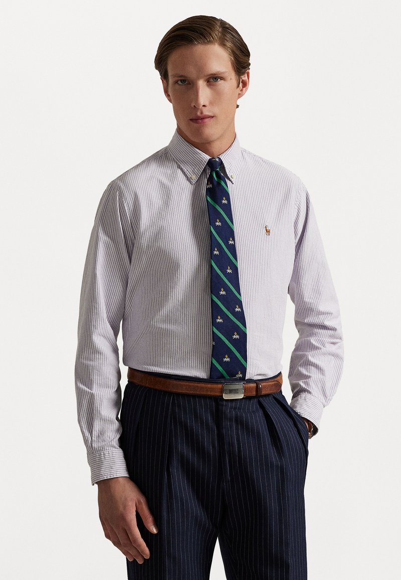Chemise pour homme à boutons, blanche à rayures bleues, accompagnée d'une cravate foncée à rayures vertes et petits motifs. Ceinture en cuir marron, pantalon à fines rayures.