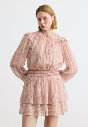 Femme portant une robe longue à manches longues rose à motifs, avec des épaules bouffantes, un décolleté en trou de serrure et une taille smockée, debout devant un fond uni.