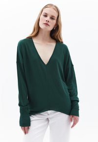 Grüner V-Ausschnitt-Pullover mit langen Ärmeln, gerippten Bündchen und lockerer Passform, kombiniert mit weißen Hosen. Weiche Textur, minimales Design.