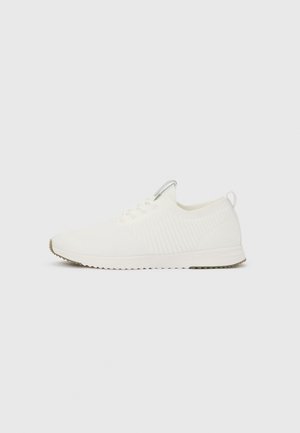Hvid lavtop atletiksneaker med snørelukning, tekstureret strikket overdel og en gummisål, vist fra siden mod en ensfarvet baggrund.