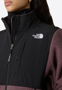 Giacca in pile nera e bordeaux con collo alto, chiusura a zip, tasca frontale e logo. Tessuto morbido con pannelli di colore a contrasto.