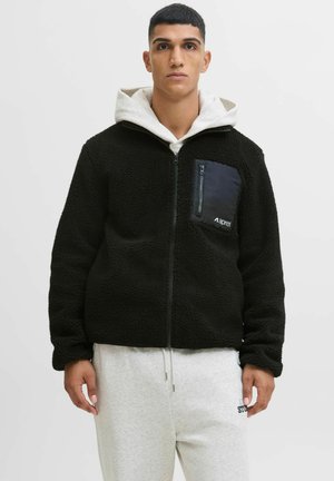 Veste en polaire noire avec une poche zippée en tissu noir, présentant un col montant et une surface texturée ; associée à un pantalon de survêtement gris clair.