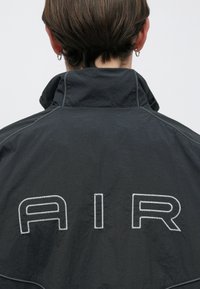 Μαύρο ελαφρύ μπουφάν με ψηλό γιακά, με λευκά ραμμένα γράμματα "AIR" στην πλάτη. Η υφή είναι λεία με ελαφριά λάμψη.