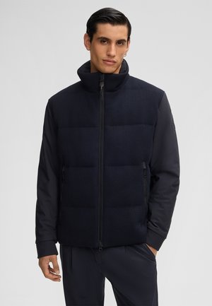 Mann trägt eine schwarze gesteppte Jacke mit Reißverschluss vorne und hat die Hände in der Nähe der Taschen vor einem schlichten hellen Hintergrund.
