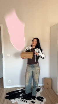 Donna con giacca bianca e nera e jeans larghi che tiene una scatola di scarpe marrone, in piedi su un tappeto bianco e nero in una stanza con pavimento in legno.