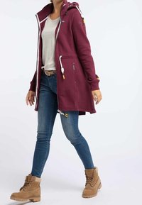 Sudadera larga color granate con capucha, cordones, dos bolsillos y textura suave, combinada con vaqueros azules y botines marrón claro.