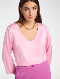 Langärmlige Bluse mit tiefem V-Ausschnitt aus glänzendem pinkfarbenem Stoff, kombiniert mit hoch taillierten rosa Hosen mit Knopfverschluss.