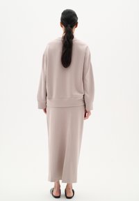 Femme aux longs cheveux bruns attachés en queue de cheval, portant un pull beige ample et une longue jupe assortie, debout de dos sur un fond blanc uni.