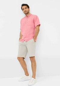 Camicia a maniche corte, con bottoni, in lino rosa chiaro con una piccola tasca frontale, abbinata a pantaloni corti beige e sneakers bianche.