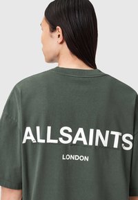 Žalios spalvos per didelis medvilninis marškinėliai, su ryškiu baltu tekstu "ALLSAINTS" ir "LONDON" ant nugaros, turi apvalų kaklą.