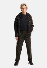 Schwarze und olivgrüne Fleecejacke mit Reißverschluss, strukturiertem Muster, kombiniert mit oliven Cargo-Hosen und dunklen Sneakers.