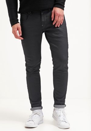 Mann trägt schwarze Slim-Fit-Hose, schwarzes Langarmshirt und weiße Sneakers mit hochgekrempelten Hosensäumen vor weißem Hintergrund.