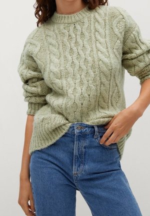Femme portant un pull torsadé vert clair à manches retroussées et un jean bleu taille haute, main posée sur la taille.