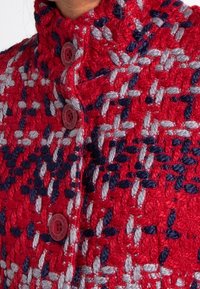 Cappotto di lana rossa con una texture a nido d'ape, decorato con un motivo intrecciato in blu navy e grigio chiaro, caratterizzato da grandi bottoni rossi e un colletto alto.
