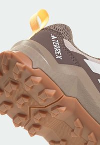 Hnědá sportovní obuv s texturovaným síťovaným svrškem, gumovou podrážkou s vícesměrným vzorkem a výraznou žlutou poutkem.