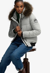 Lys grå pufferjakke med en faux fur hætte, lynlåslukning og trykknaplukning samt et mærke på ærmet; parret med en grøn sweater og blå jeans.