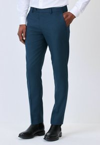 Tmavě modré kalhoty vyrobené z hladkého materiálu, s úzkým střihem, bočními kapsami a hladkým předním designem, nošené s černými kotníkovými botami.