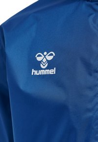 Blå jacka gjord av ett slätt, glänsande material med en vit Hummel-logotyp på övre vänstra sidan. Designen är enkel utan ytterligare dekorationer.