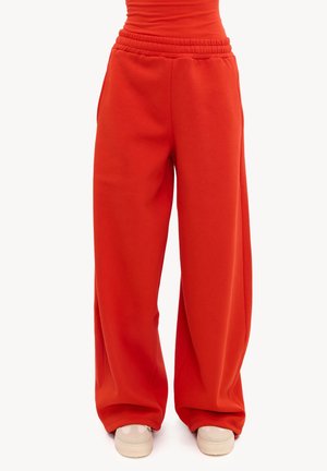 Pantalon rouge à jambes larges avec une taille élastique porté avec des baskets blanches et un haut orange, montré de la taille aux pieds.