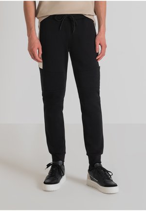 Pantalones de chándal negros con cintura ajustable mediante cordón, con paneles texturizados. Combinados con zapatillas negras y blancas.