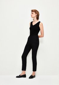 Svart ärmlös topp som matchas med h�ögmidjade, avsmalnande svarta byxor. Byxorna har en slits vid fållen. Modellen bär svarta loafers.