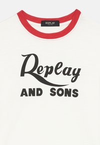 Fehér pamut póló piros bordázott nyakkal. Jól látható fekete nyomtatás található rajta, amelyen a "Replay AND SONS" felirat különböző betűtípusokban olvasható.