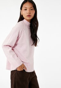 Maglione a maglia rosa chiaro con un collo alto a costine, orlo a costine e una vestibilità rilassata, indossato con pantaloni marroni; texture morbida e design informale.