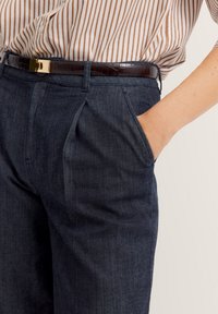 Dunkelblaue Hose mit Falten, seitlichen Taschen und glatter Textur. Kombiniert mit einem braunen Gürtel und einem weiß gestreiften Hemd.