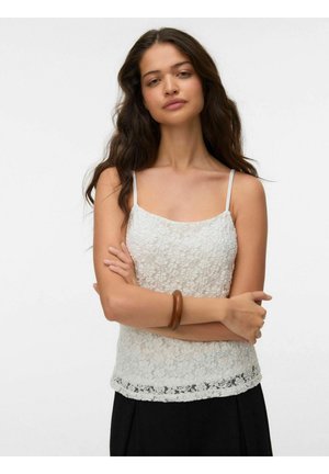 Kvinde med langt mørkt hår iført en hvid blonder topp med spaghetti stropper og sorte bukser, med armene krydset, med et træ armbånd på håndleddet.