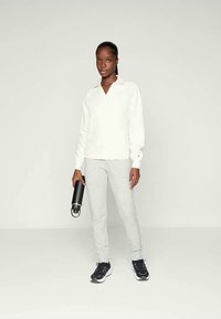 Maglione bianco a maniche lunghe con scollatura a V, abbinato a pantaloni della tuta grigi chiari con motivo e scarpe da ginnastica nere. Tiene in mano una bottiglia d'acqua nera.