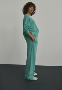 Türkiser Sweatshirt und passende Hose aus weichem Material. Lockere Passform mit seitlichen Taschen, gerippten Bündchen und dezenten Nahtdetails. Silberne Sneaker.