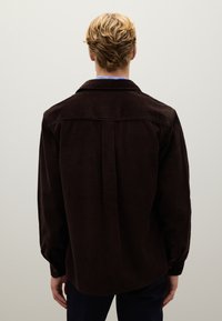 Donkerbruine corduroy overhemd met een rugstuk en lange mouwen, voorzien van knoopmanchetten en verticale ribbelstructuur.