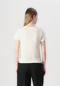 T-shirt a maniche corte di colore crema con scollatura tonda. Presenta una cucitura centrale lungo la schiena e una texture morbida. Indossato con pantaloni neri.