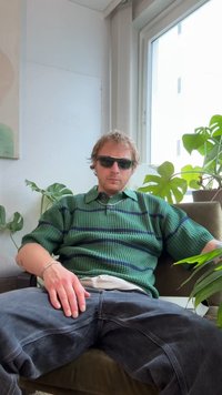 Πράσινο πόλο πουκάμισο με μπλε ρίγες, μαύρα γυαλιά ηλίου και σκούρο τζιν παντελόνι· καθισμένος σε πολυθρόνα με φυτά. Ουδέτερο φόντο.