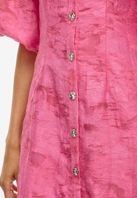 Abito rosa strutturato con una chiusura frontale a bottoni e accenti floreali metallici. Il tessuto presenta un motivo sottile ed ha un aspetto leggero.