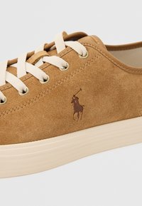 Tan mockasinsneakers med beige snören och ögon av guldton. Har en beige sula och en liten, präglad polo-logotyp på sidan.