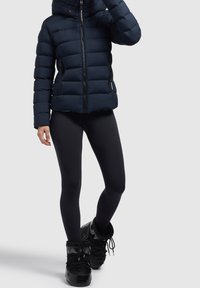 Marineblauwe donzen jas met kap, ritssluiting en zijzakken, gecombineerd met zwarte leggings en zwarte winterlaarzen. Gladde textuur.