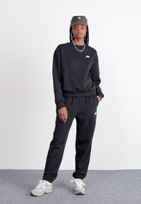 Ensemble de sweat-shirt et pantalon noir en tissu doux, avec un petit logo sur la poitrine. Porté avec une casquette grise et des baskets blanches.